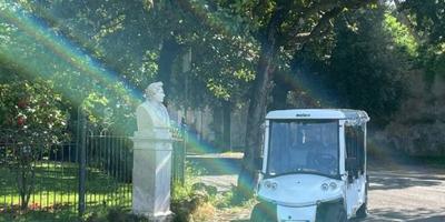Un pequeño carrito de golf eléctrico pasando junto a un busto de mármol en un sendero sombreado del parque en Roma